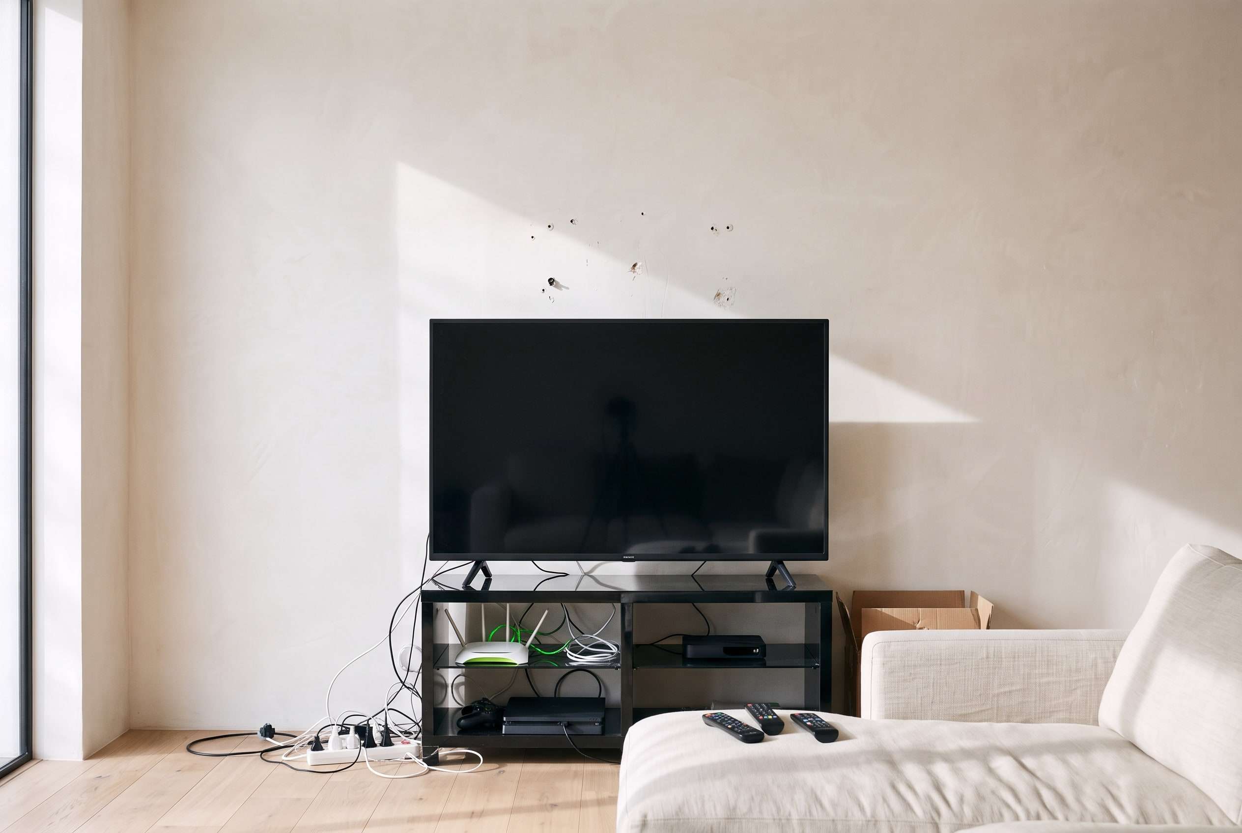 Voor: een televisie op een los tv-meubel met kabels, een stekkerdoos, een router en afstandsbedieningen, en boorgaten in de muur.