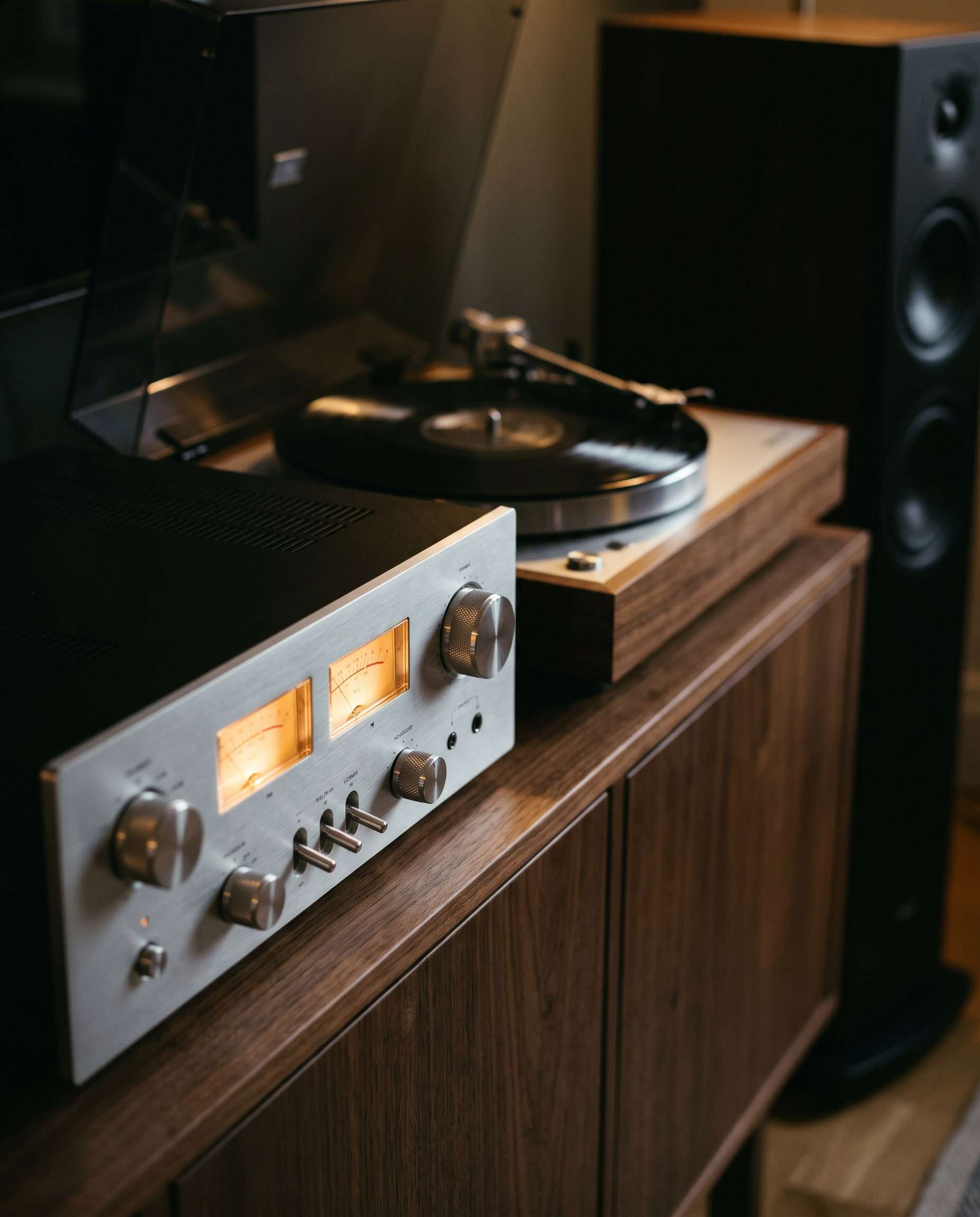 Sfeerbeeld: een zilverkleurige versterker met oplichtende VU-meters naast een draaiende platenspeler op een notenhouten kast.